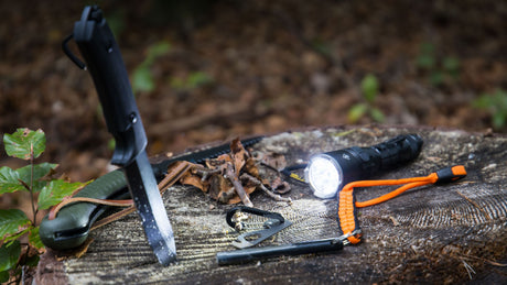 Eine eingeschaltende Tschenlampe, ein Messer, eine Handsäge und ein Feuerstahl liegen auf einem Baumstumpf