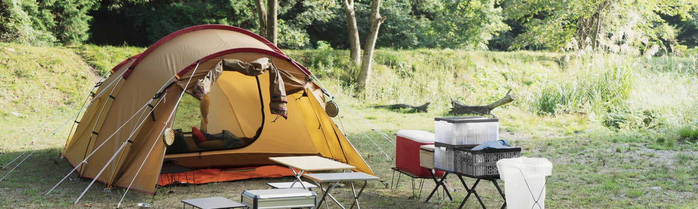 Ein Campingzelt steht aufgebaut zusammen mit Camping-tischen und Stühlen auf einer Wiese: Campingausrüstung mieten