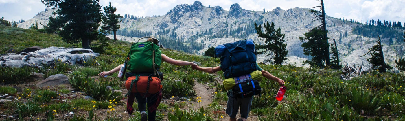 Zwei Wanderer rennen Händchen haltend in Richtung Bergpanorama: Tourenrucksack mieten